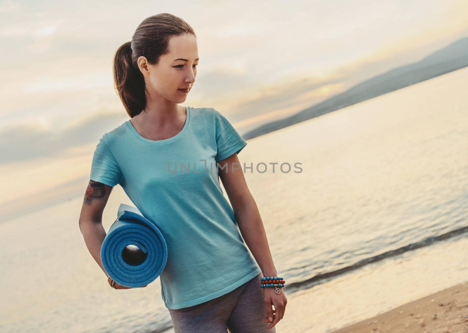 Young beautiful woman with yoga mat standing on beach in summer and ready for workout.