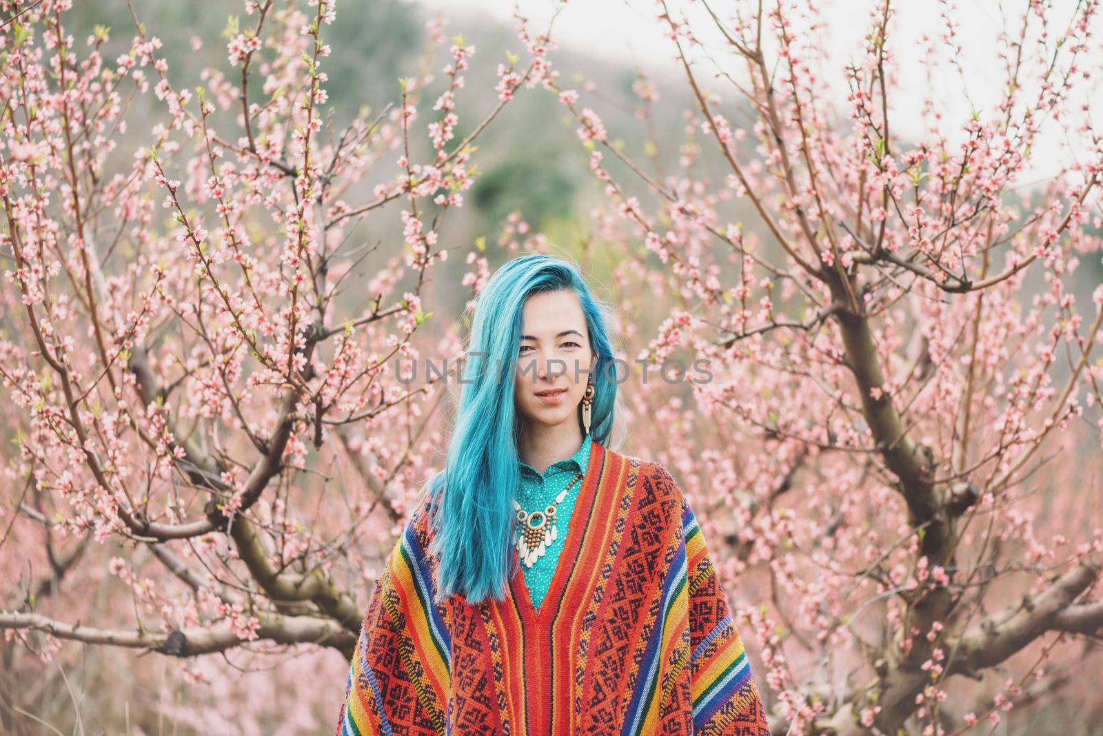 Beautiful young woman with blue hair wearing in poncho walking among the flowering pink trees in spring.