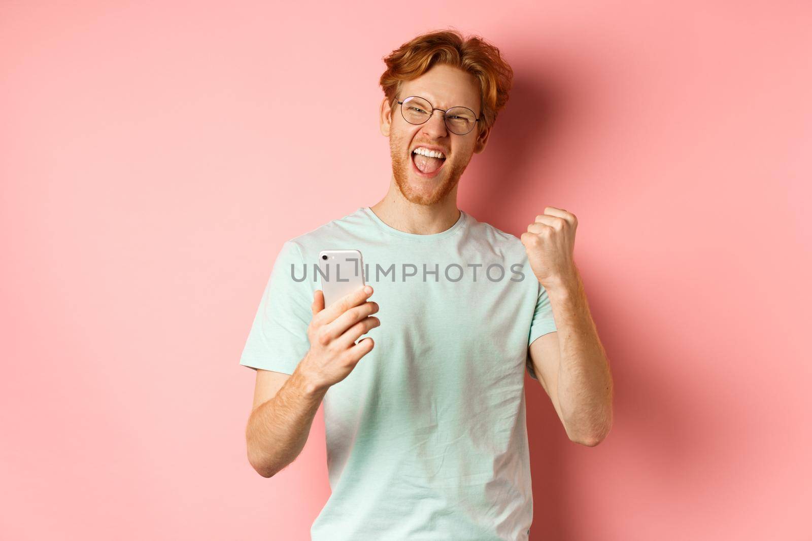 Happy redhead guy in glasses and t-shirt winning online prize, shouting yes with joy and satisfaction, holding smartphone and making fist pump, pink background by Benzoix