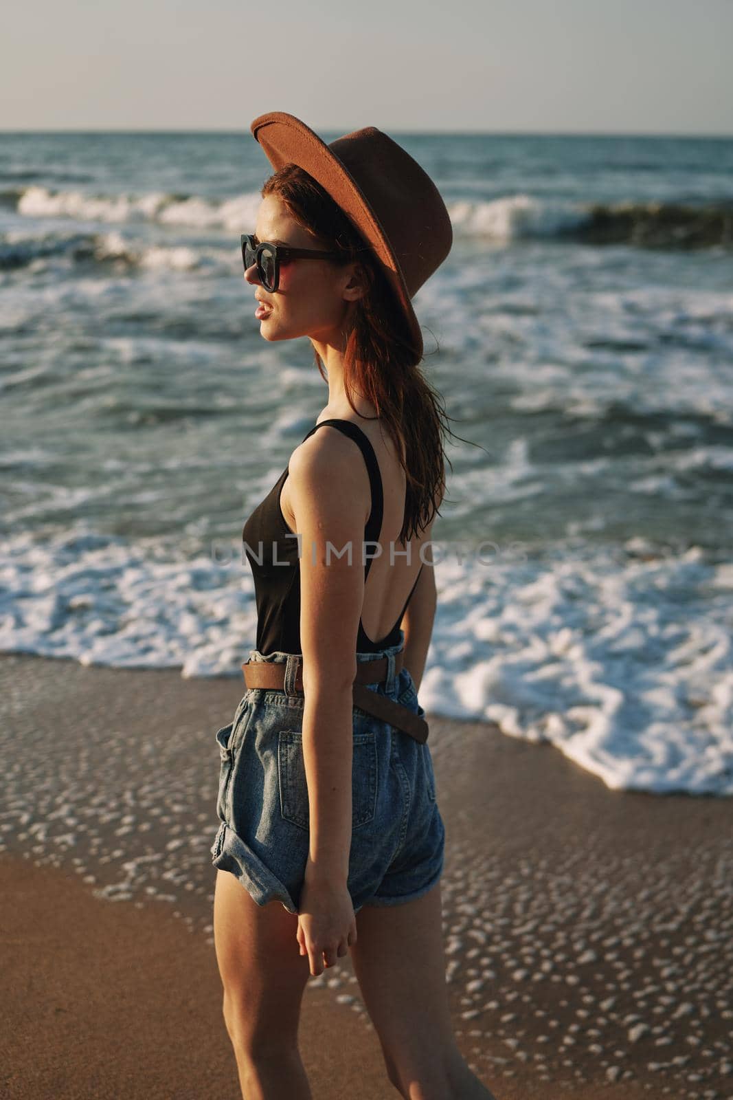 cheerful woman in sunglasses and a hat walks along the ocean by Vichizh