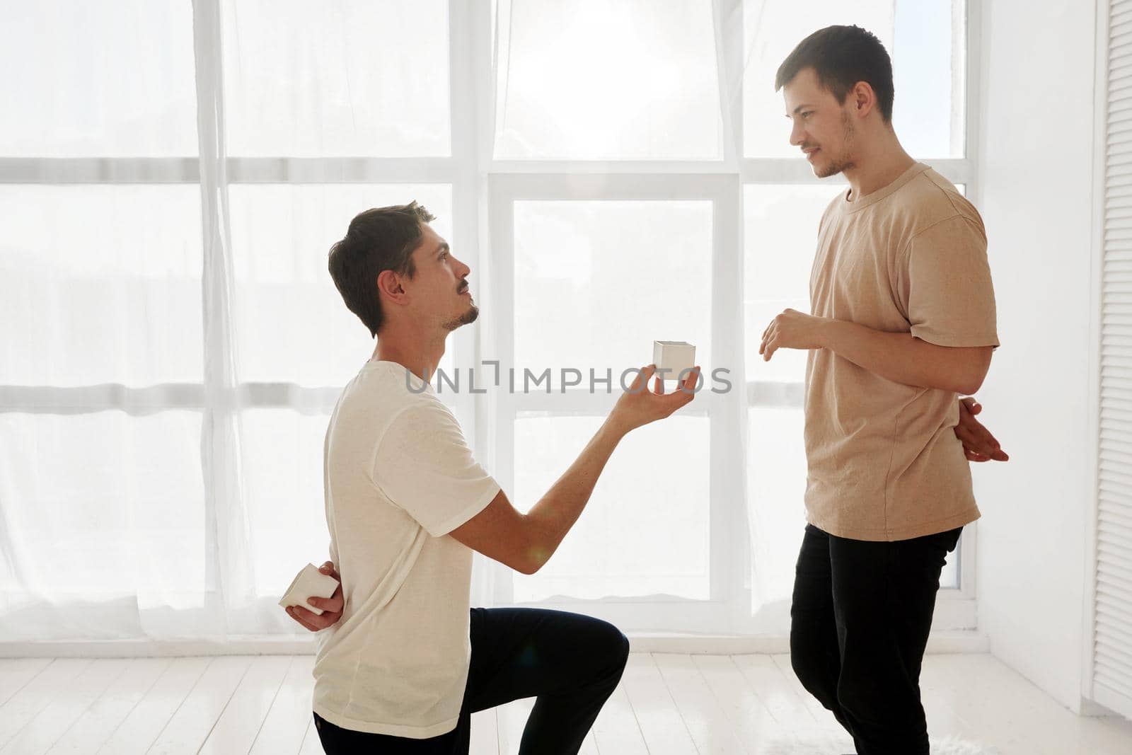 Side view of happy gay man kneeling and proposing to loving man against white window