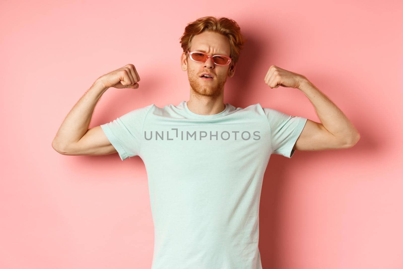 Confident young man with red hair, wearing summer sunglasses and t-shirt, showing strong and fit body muscles, flex biceps and staring cool at camera, pink background by Benzoix