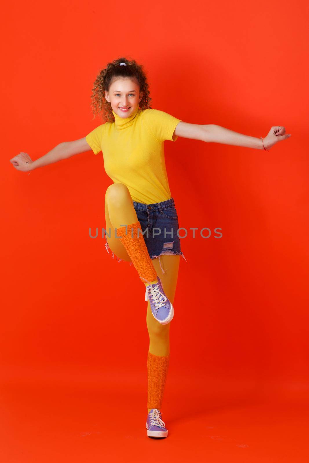Girl in sports outfit jumping on one leg. Joyful girl with ponytail looking stylish in turtleneck blouse, leggins, shorts and socks gaiters jumping with her arms outstretched against red background