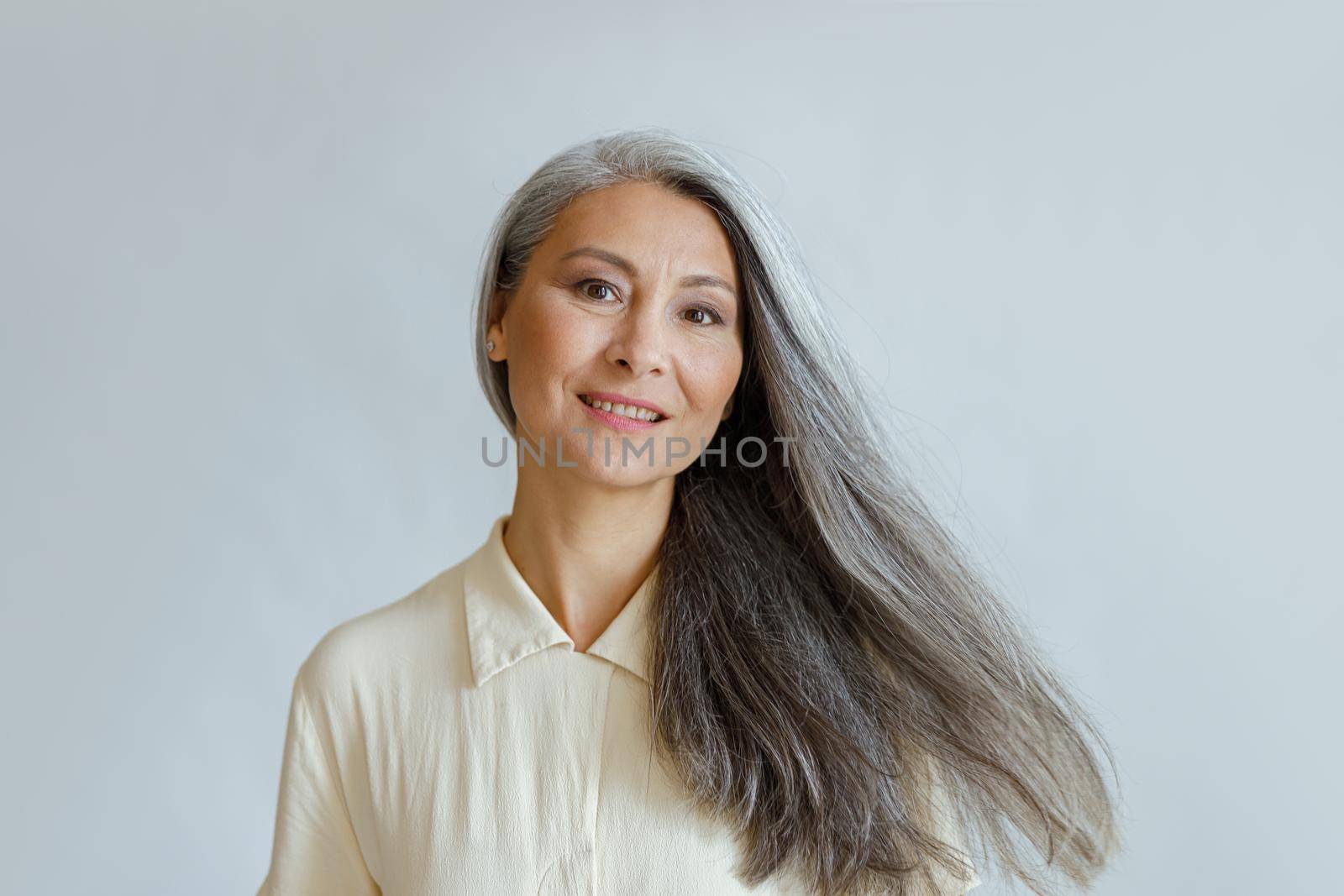 Smiling middle aged Asian woman with flowing grey hair stands on light background by Yaroslav_astakhov