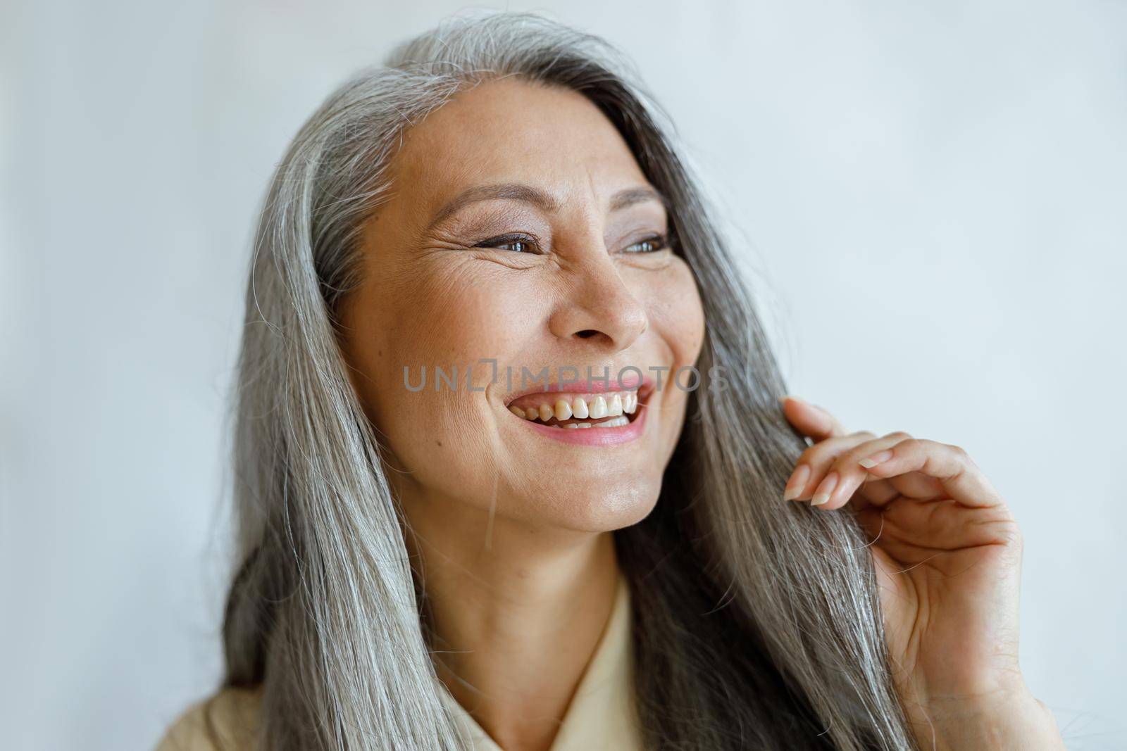 Joyful middle aged Asian woman with natural grey hair smiles on light background in studio closeup. Mature beauty lifestyle