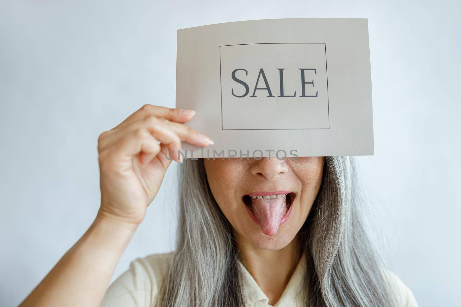 Playful middle aged lady with straight silver hair holds card with word Sale covering face and shows tongue on light background in studio closeup