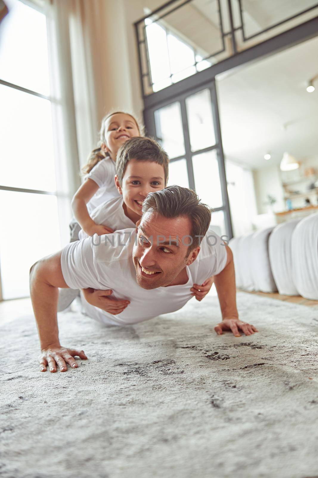 Cheerful sporty man is doing push ups in living room with happy son and daughter on back