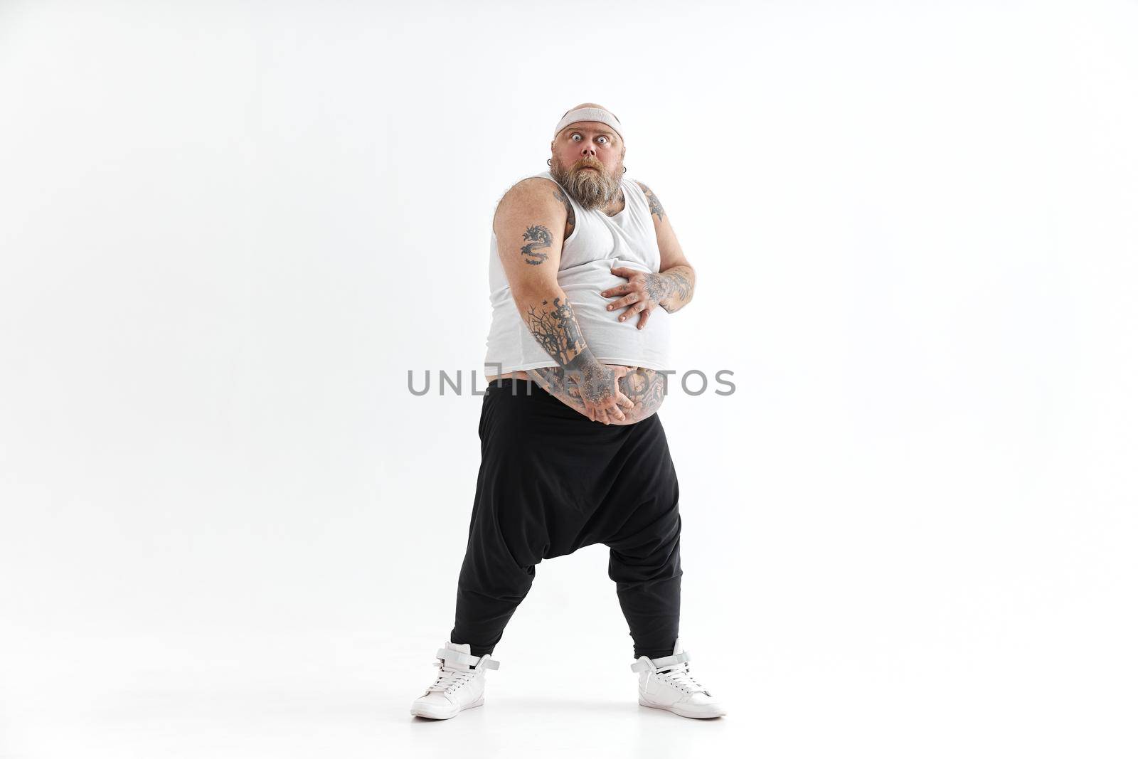 Happy fat man with big belly and tattoes in sports wear is holding his stomach by Yaroslav_astakhov