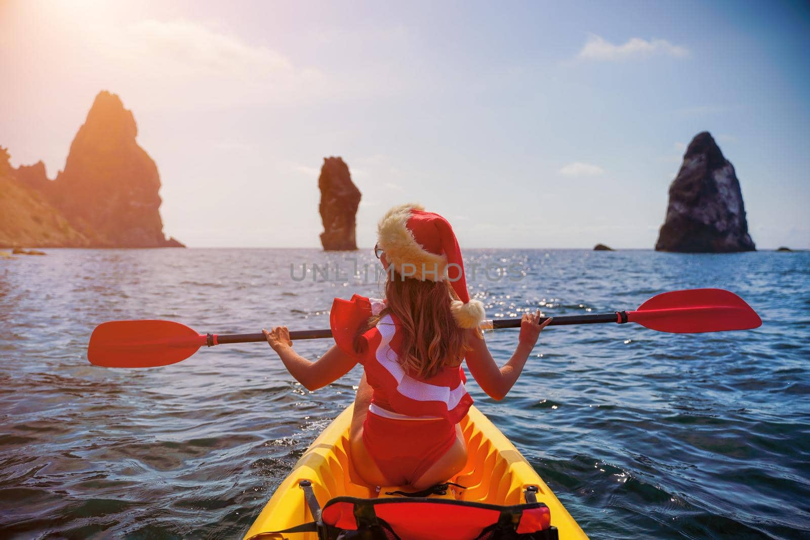 Young brunette woman in red swimsuit and Santa hat, swimming on kayak around basalt rocks like in Iceland. Back view. Christmas and travel concept