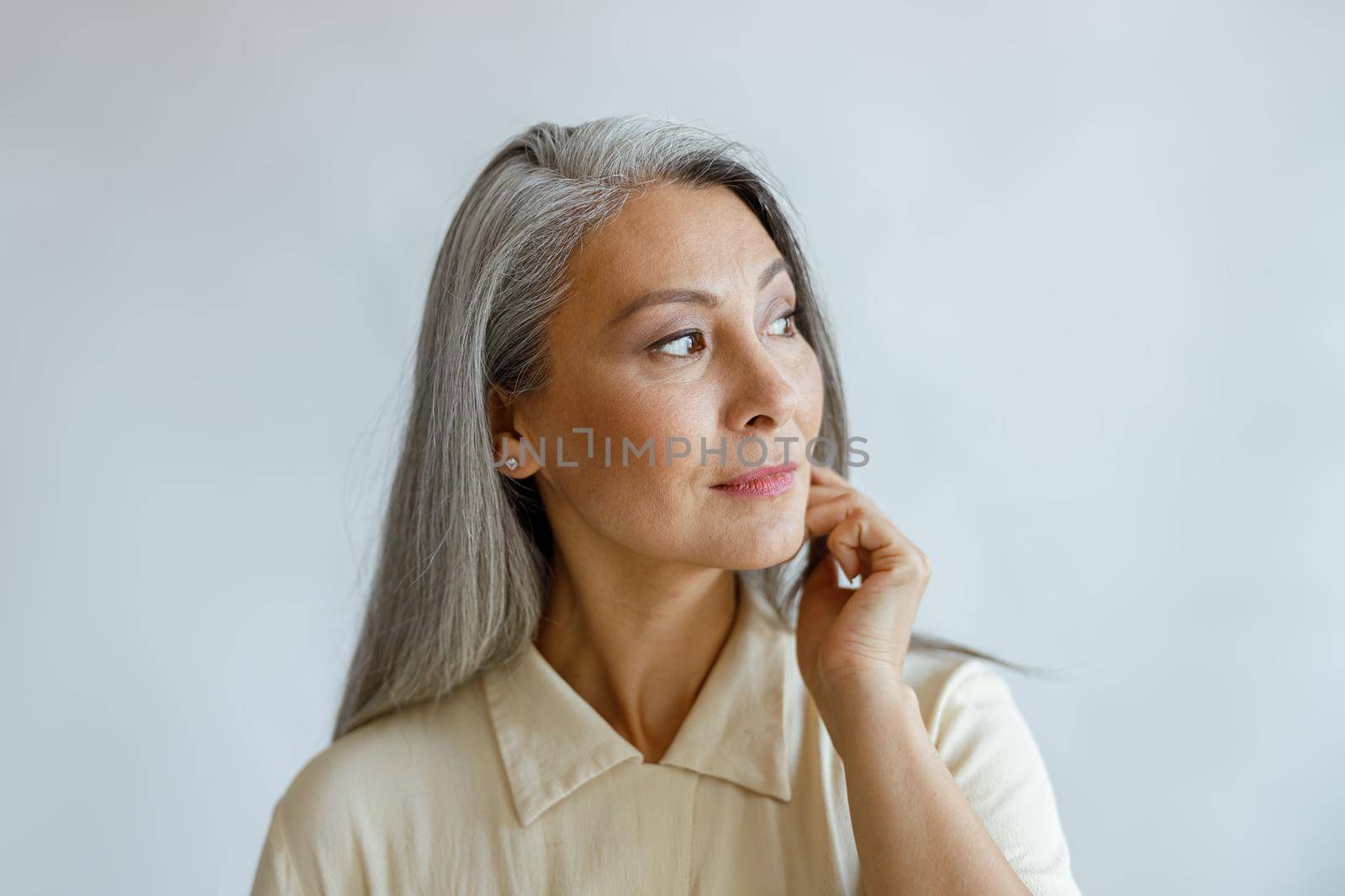 Thoughtful grey haired woman in stylish shirt looks aside on light background in studio. Mature beauty lifestyle