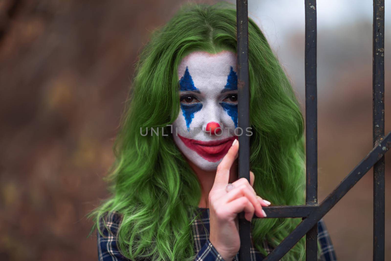 Close-up portrait of a greenhaired girl in chekered dress with joker makeup on a blurry brown background.