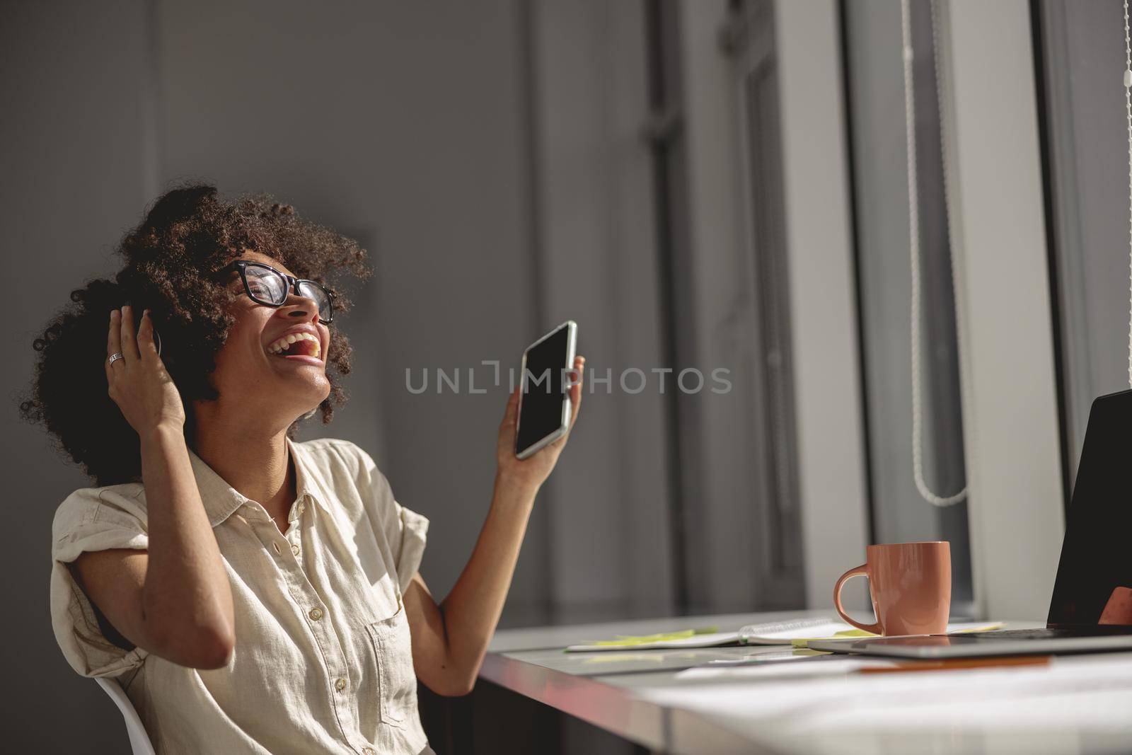 Happy Afro American female sitting in the coworking headphones and holding mobile phone