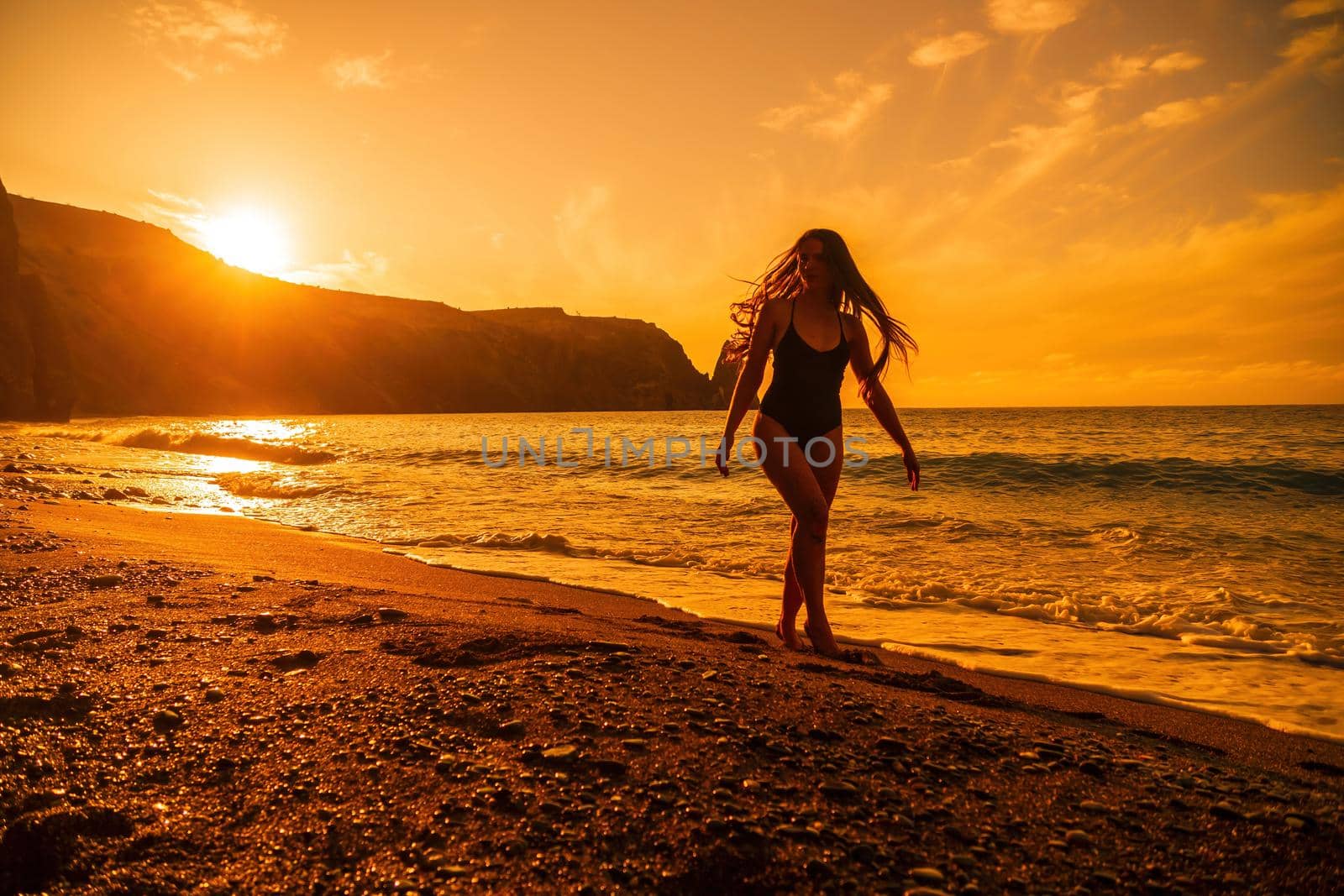 Selective focus. Happy carefree sensual woman with long hair in black swimwear posing at sunset beach. Silhouette of young beautiful playful positive woman outdoor. Summer vacation and trip concept.