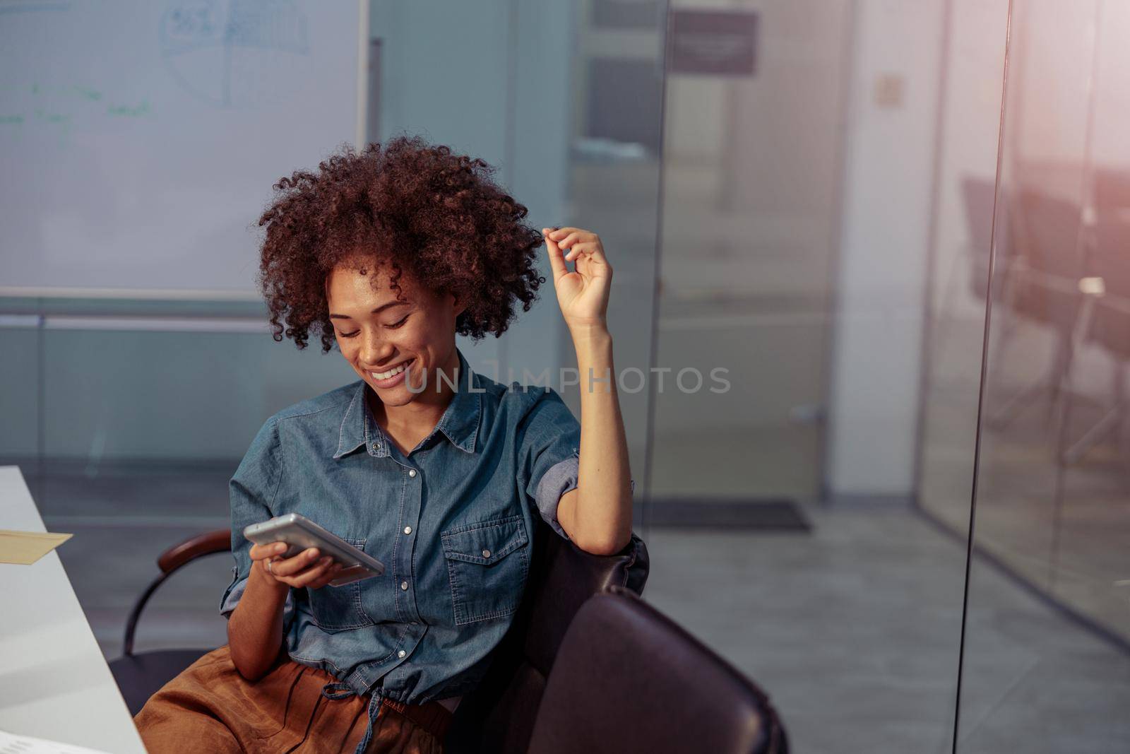 Smiling Afro American lady sitting in meeting room in the office while holding smartphone. Copy space