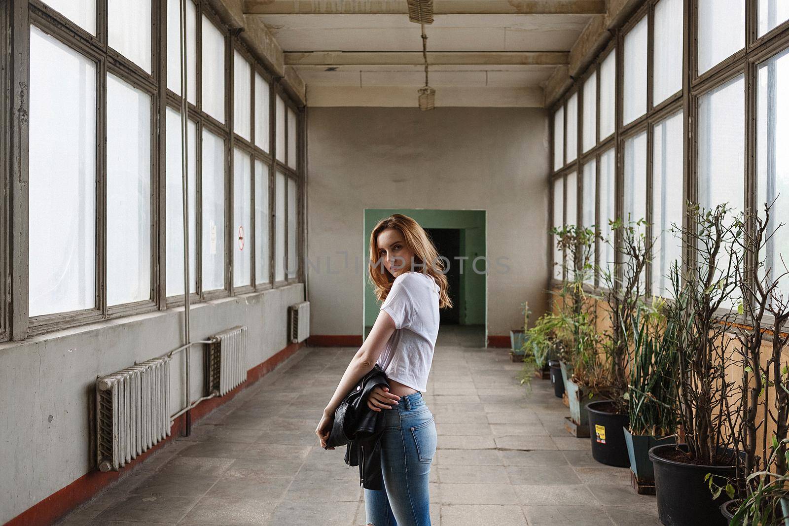 portrait of young caucasian woman standing in white t-shirt, blue ripped jeans at coridor with windows, radiators and houseplants. pretty lady stands in centre of old room