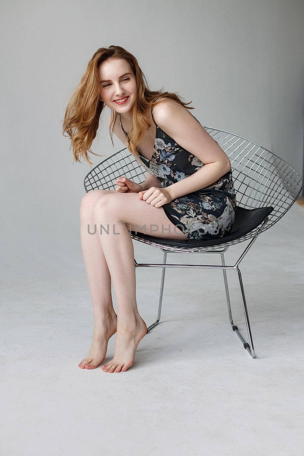 Beautiful young woman in motley dress, indoors portrait of cute smiling model. caucasian skinny female with long brown hair sitting in chair on white background. natural pretty lady posing at home