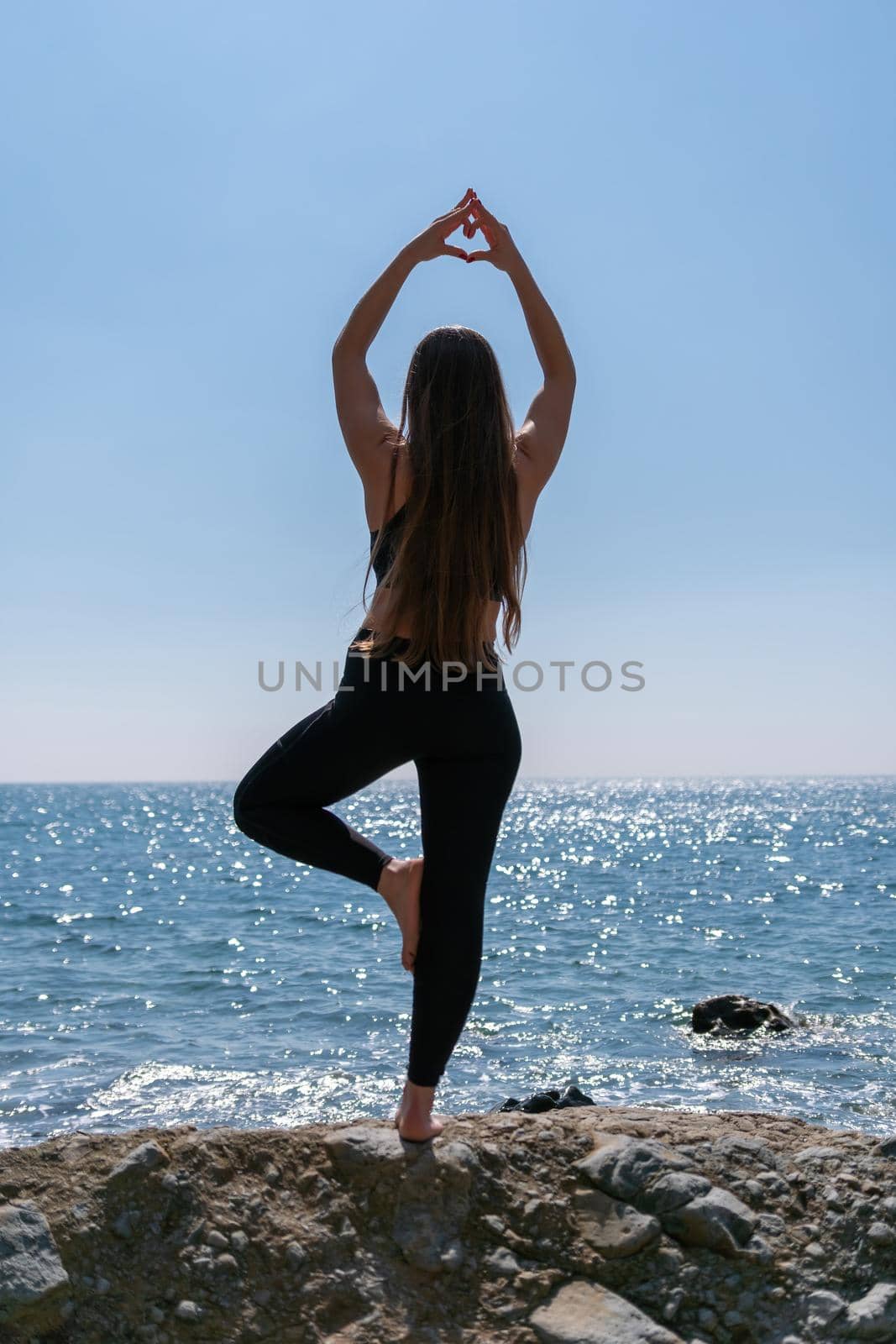 Woman makes heart with hands on beach. Young woman with long hair, fitness instructor, stretching before pilates, on a yoga mat near the sea on a sunny day, female fitness yoga routine concept by panophotograph