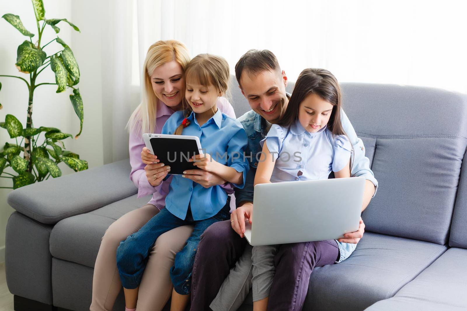 Happy family of four parents and cute little kids children enjoy using the tablet, watching cartoons, make internet video call or shopping online looking at computer screen sit together at home by Andelov13