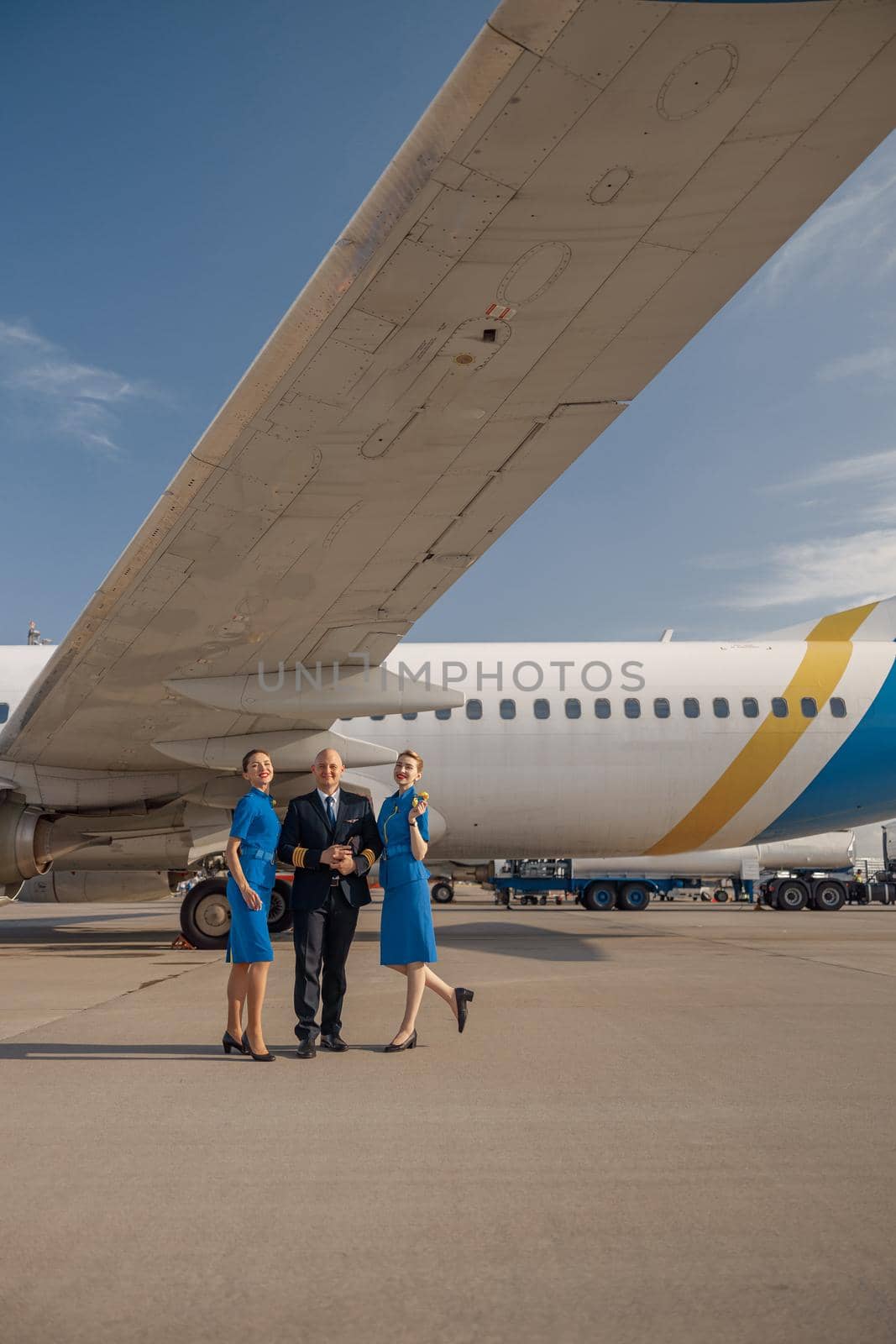 Full length shot of pilot standing together with two stewardesses in bright blue uniform in front of an airplane on a sunny day by Yaroslav_astakhov