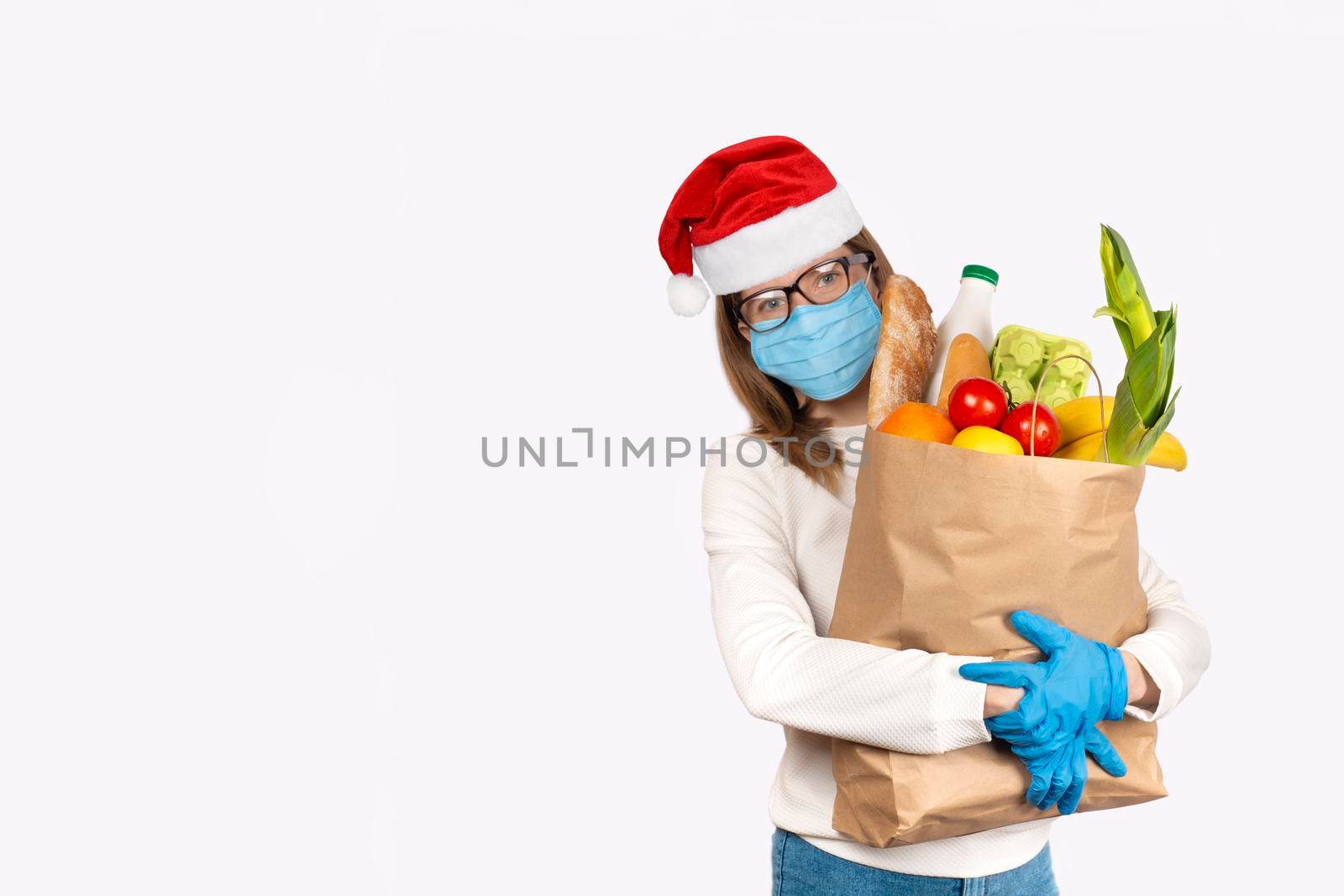 Safe food delivery or donation concept. Food delivery or shopping during coronavirus quarantine. Young woman in disposable gloves, face mask and Santa hat holding Box with food ingredients
