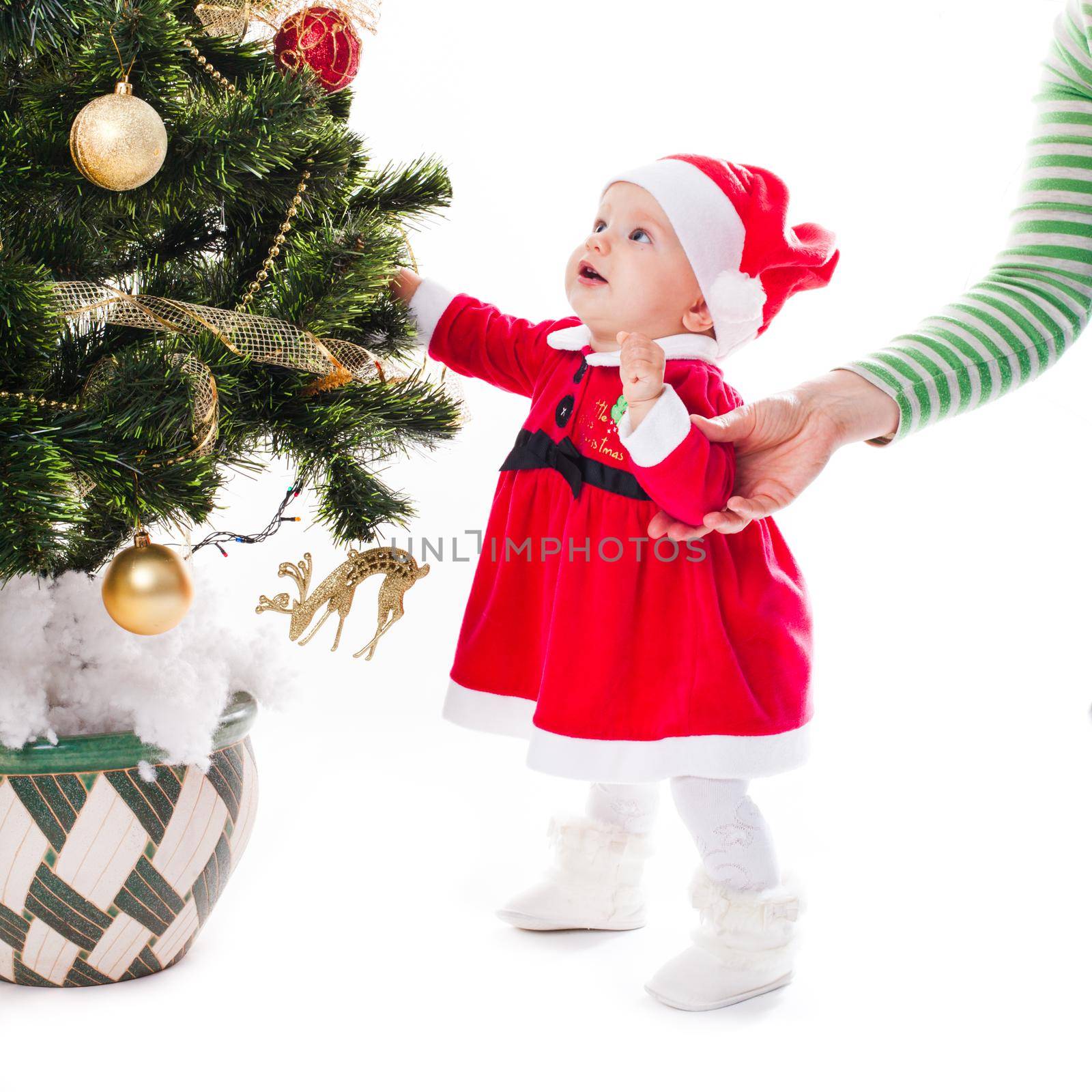 Santa baby girl in dress near Christmas tree with mother's hand - my first holiday