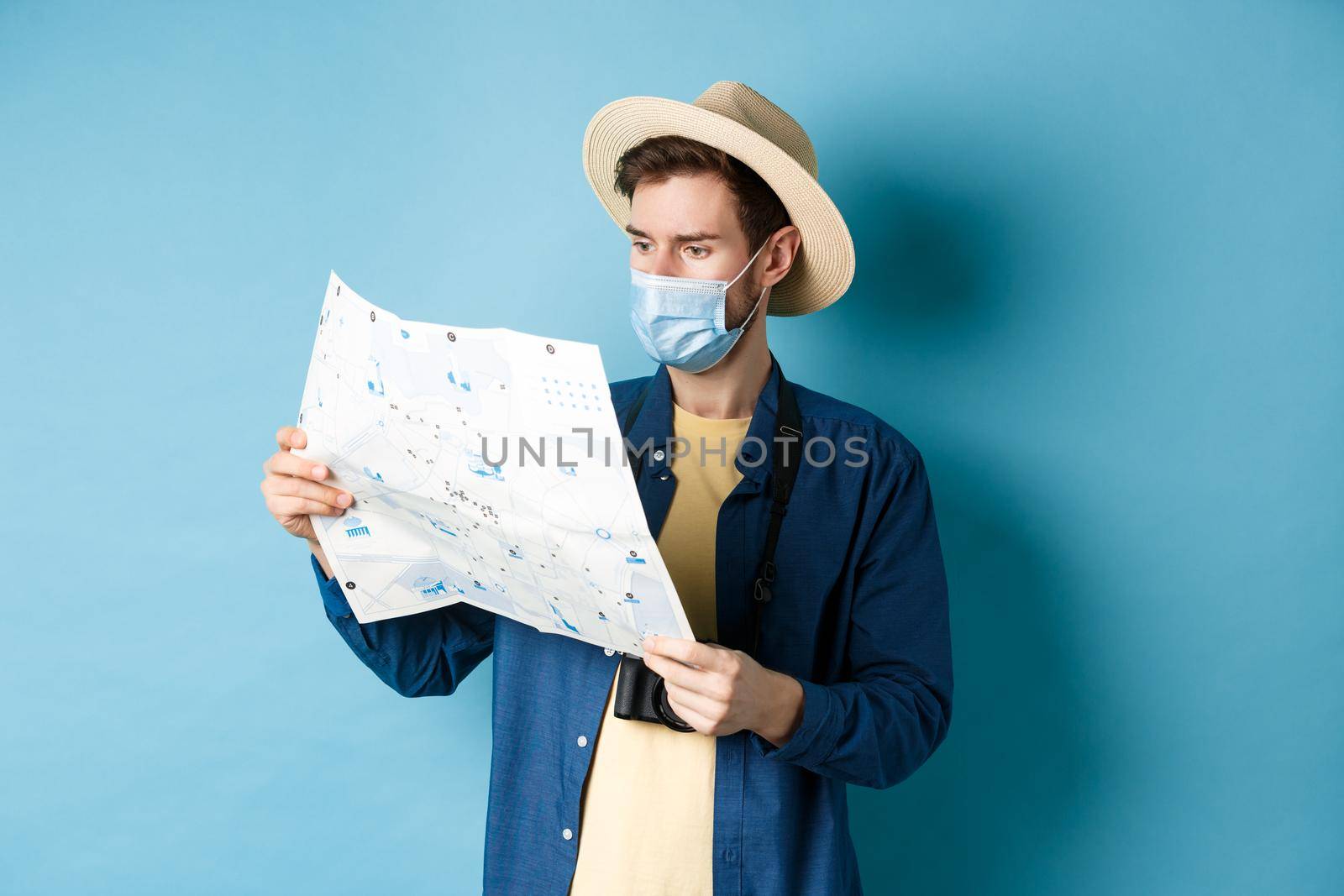 Covid-19, pandemic and travel concept. Handsome young guy in face mask and summer hat looking at tourist map, travelling on vacation, standing on blue background by Benzoix