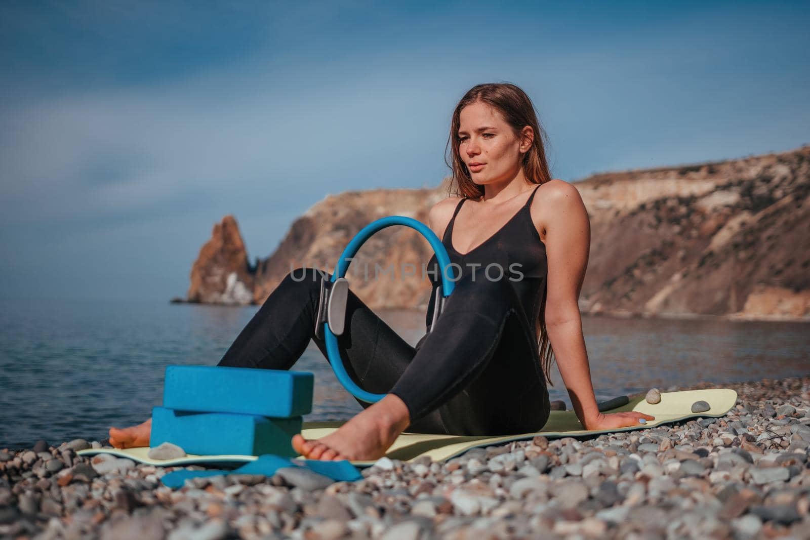 Young woman with long hair, fitness instructor in black Sportswear Leggings and Tops, stretching on a yoga mat with magic pilates ring near the sea on a sunny day, female fitness yoga routine concept.