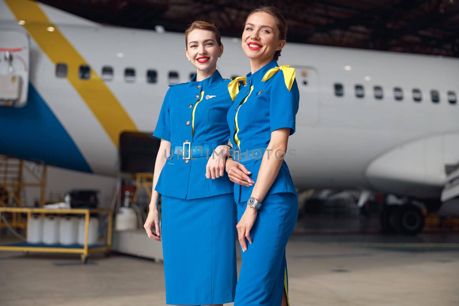 Two air stewardesses in stylish blue uniform smiling at camera, standing together in front of passenger aircraft in hangar at the airport. Occupation concept