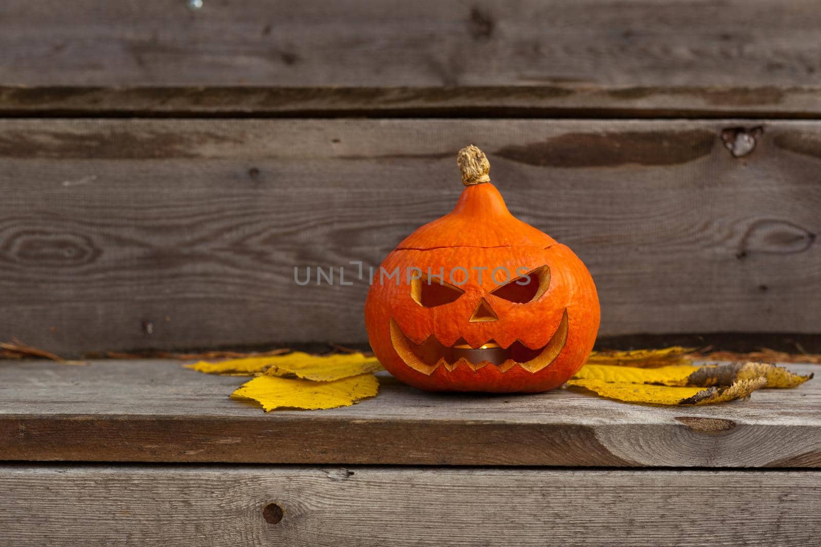 halloween scary pumpkin with eyes and mouth and yellow leaves on wooden background. Autumn Carved funny handmade Pumpkin