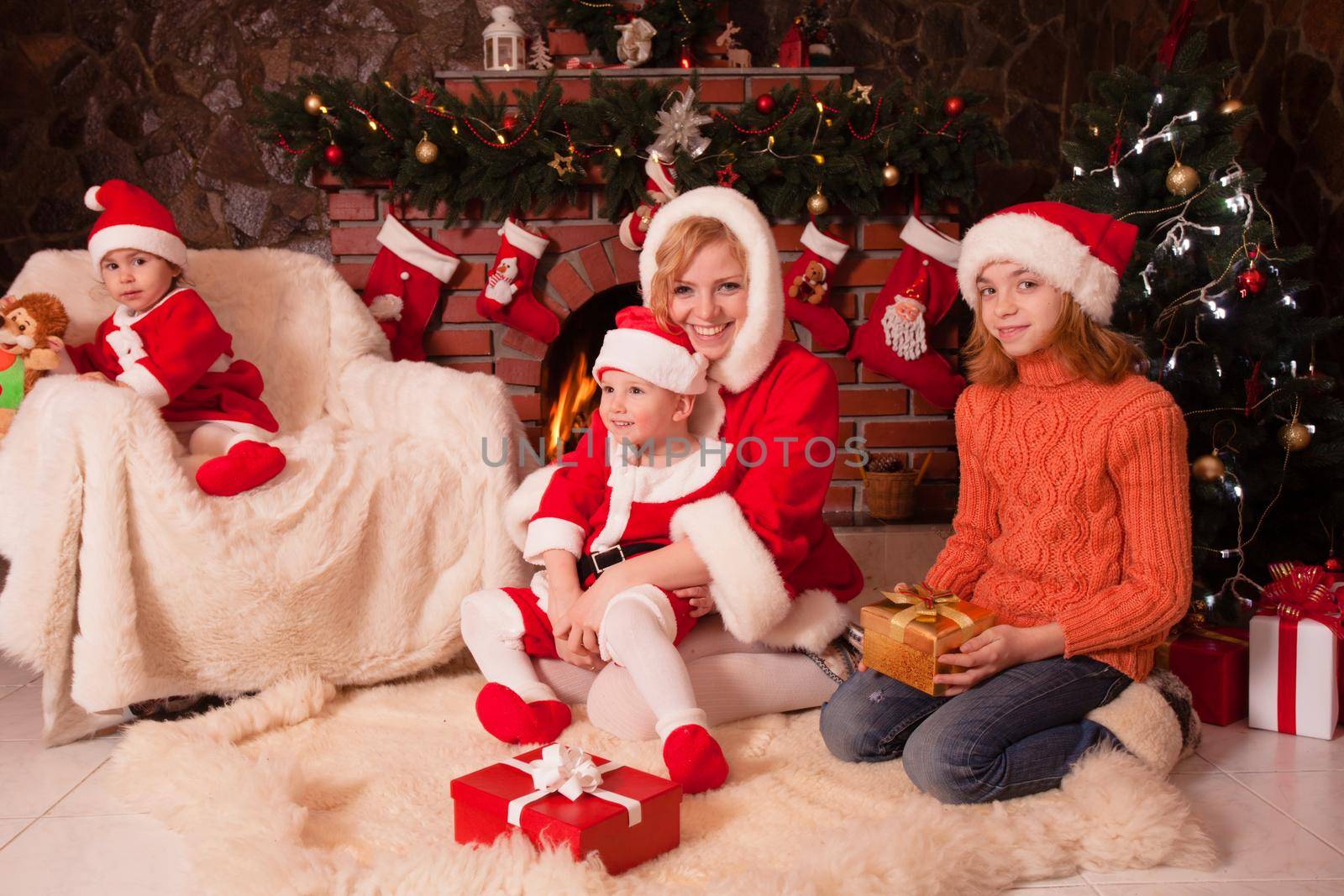 Mother and children are sitting near fireplace and christmas tree with gift boxes. Brother and sister in santa suits