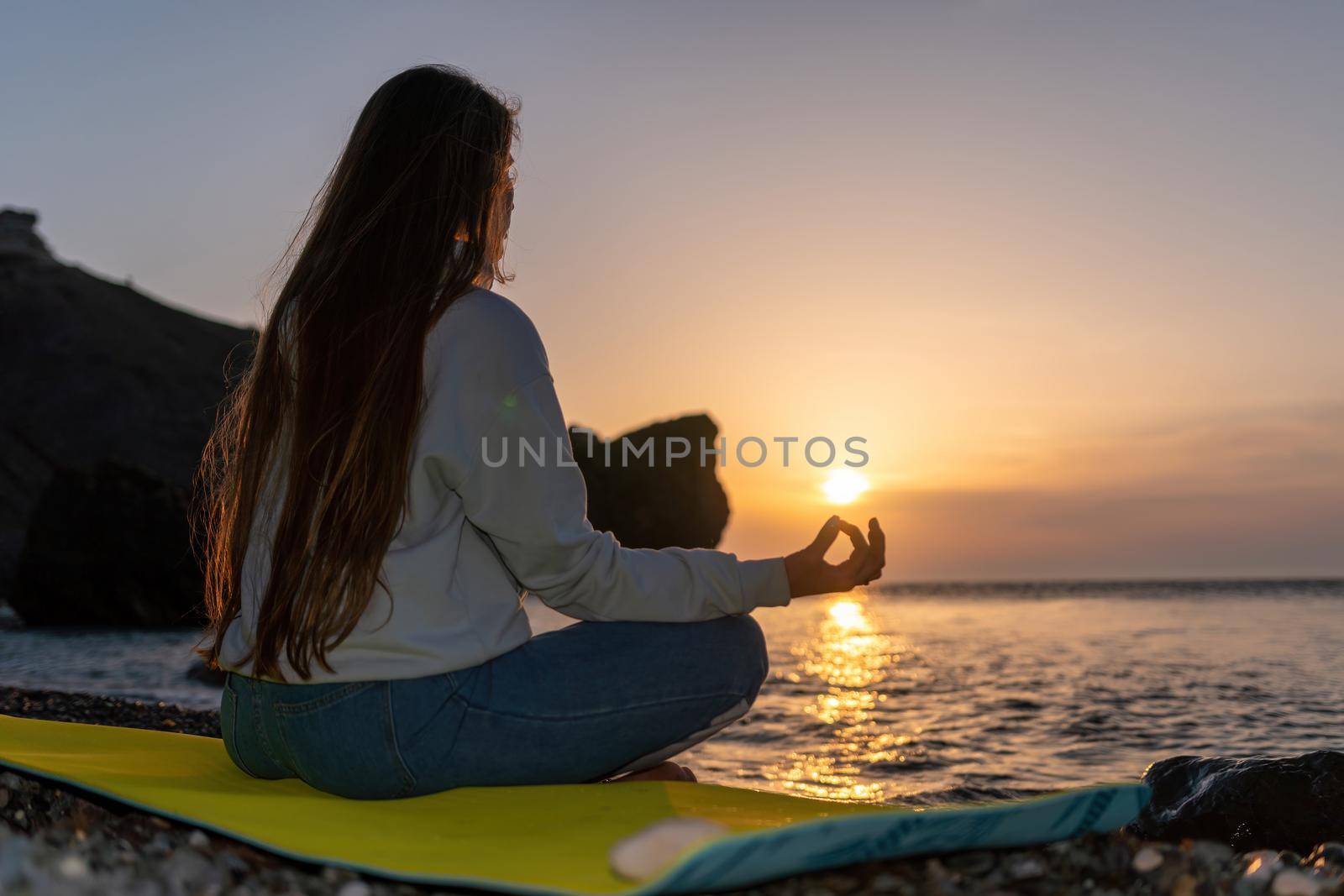 silhoute of Young woman with long hair in blue jeans and sportswear practicing outdoors on yoga mat by the sea on a sunset. Women's yoga fitness routine. Healthy lifestyle, harmony and meditation by panophotograph