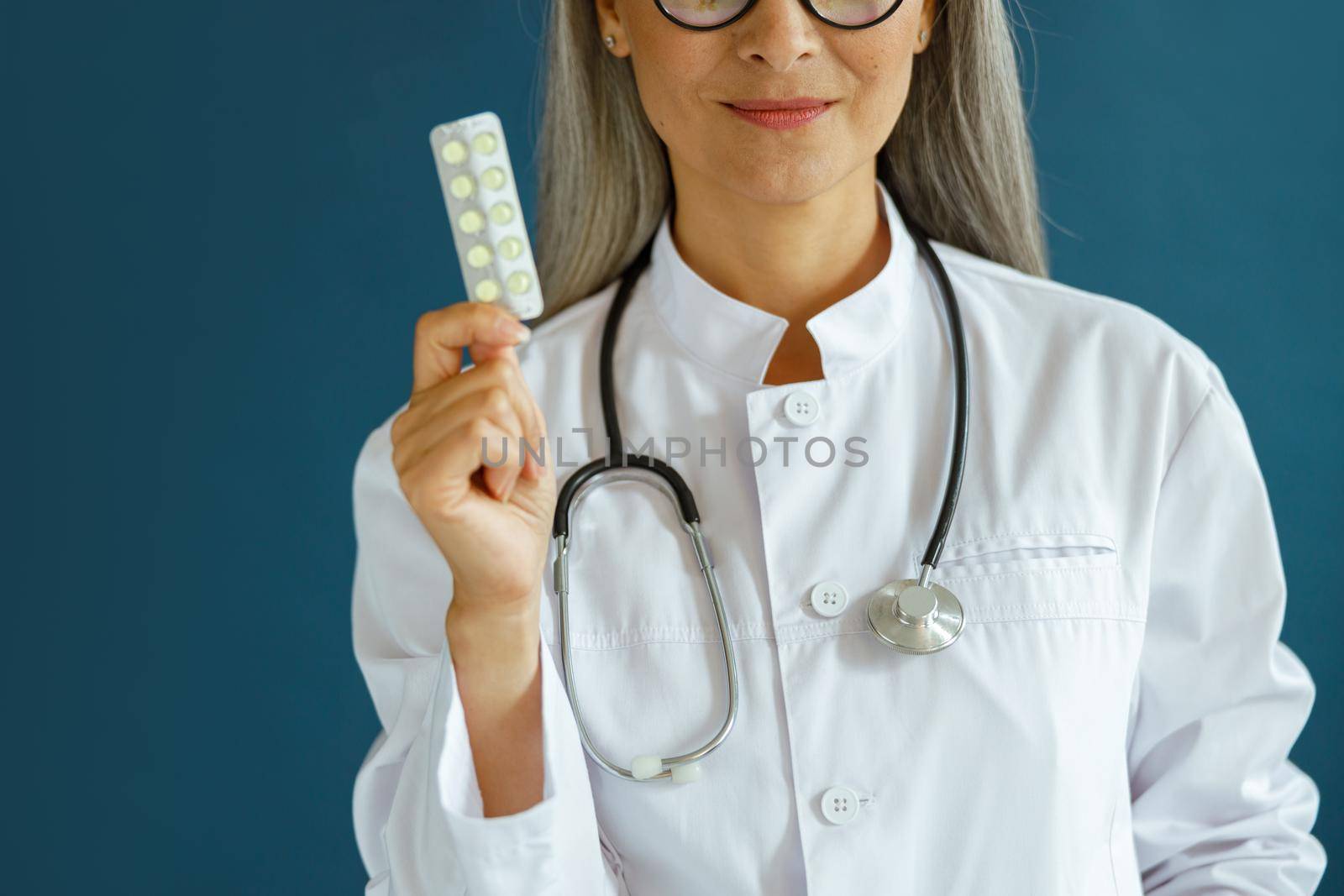 Happy mature lady therapist in white coat shows pills standing on blue background in studio closeup. Professional medical staff