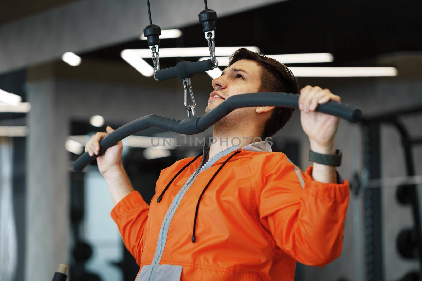 Young man in orange windbreaker working out training back muscles in the gym