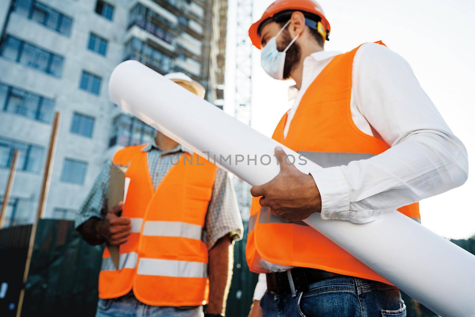 Two men in workwear and medical masks working with blueprints on object, close up