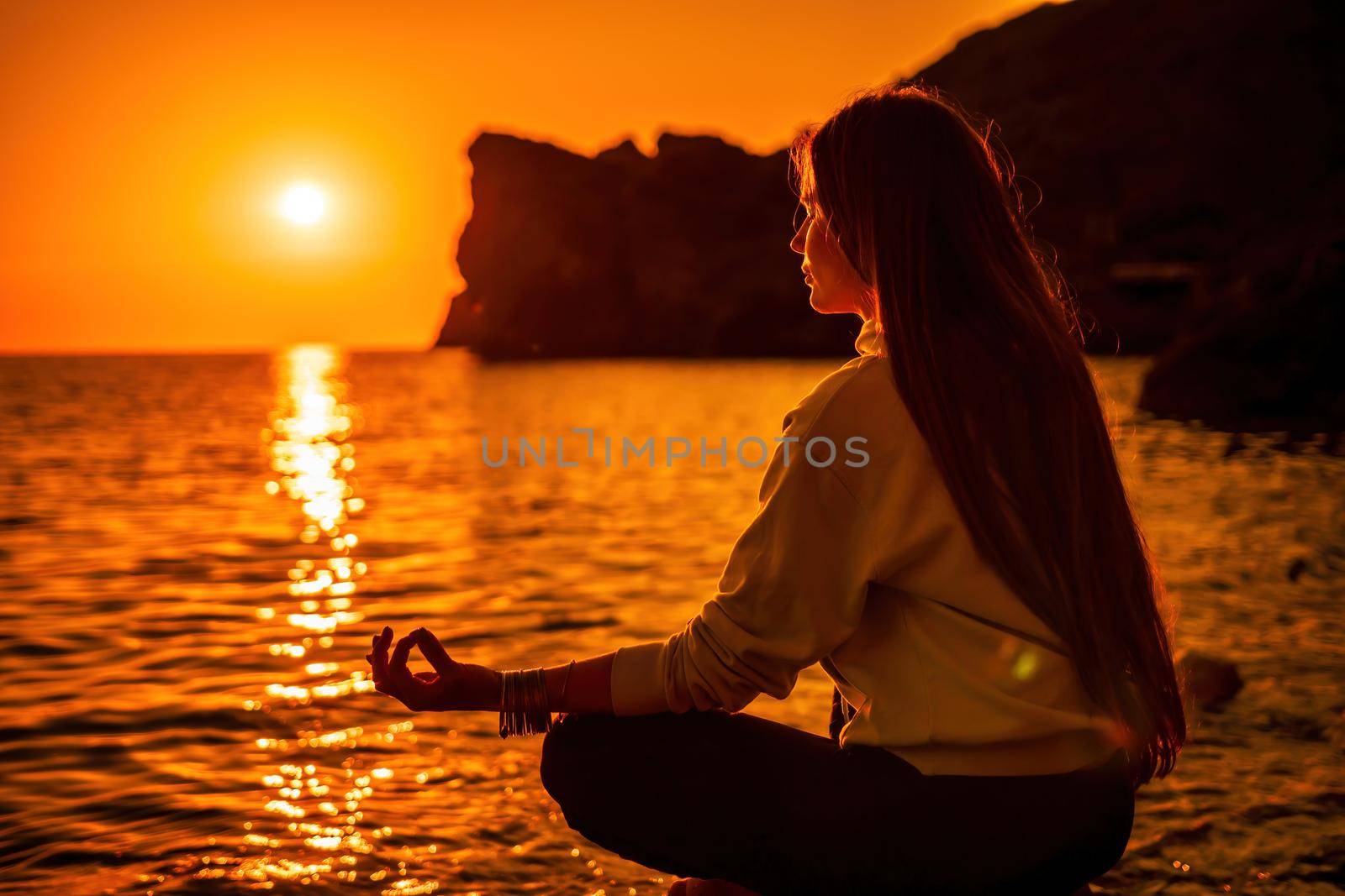 Young woman with long hair in sportswear and boho style braclets practicing outdoors on yoga mat by the sea on a sunset. Women's yoga fitness routine. Healthy lifestyle, harmony and meditation by panophotograph