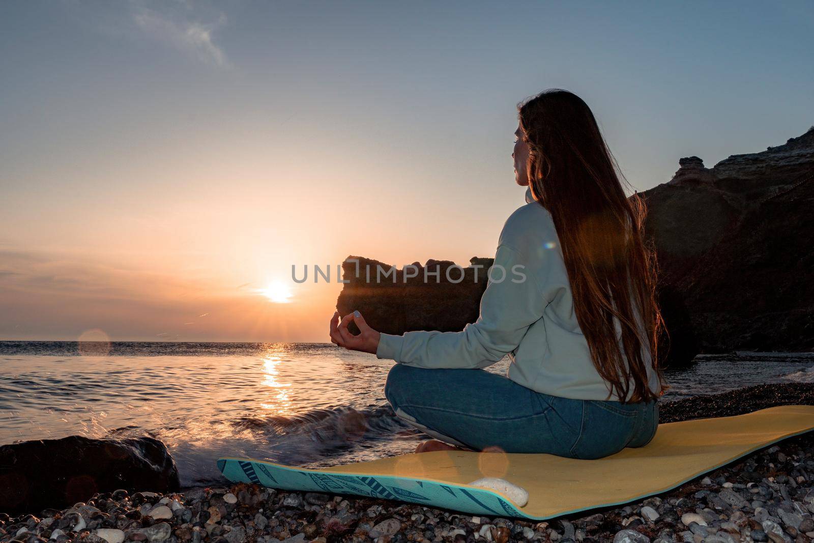 silhoute of Young woman with long hair in blue jeans and sportswear practicing outdoors on yoga mat by the sea on a sunset. Women's yoga fitness routine. Healthy lifestyle, harmony and meditation by panophotograph