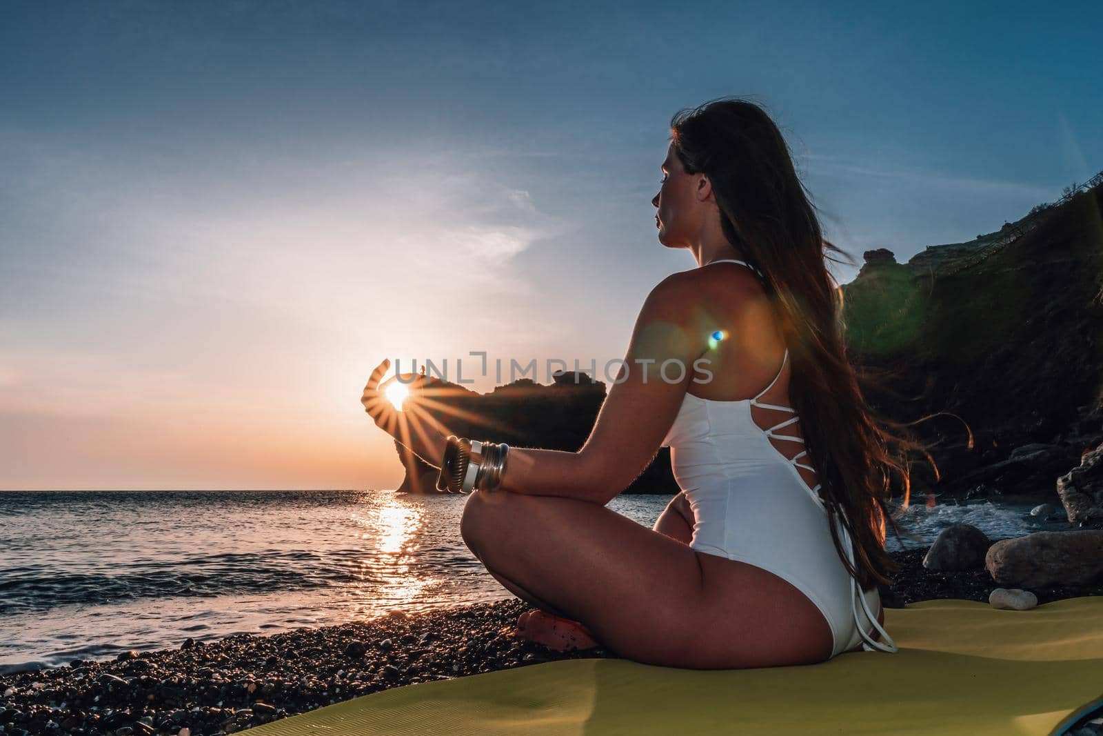 Young woman in swimsuit with long hair practicing stretching outdoors on yoga mat by the sea on a sunny day. Women's yoga fitness pilates routine. Healthy lifestyle, harmony and meditation concept.