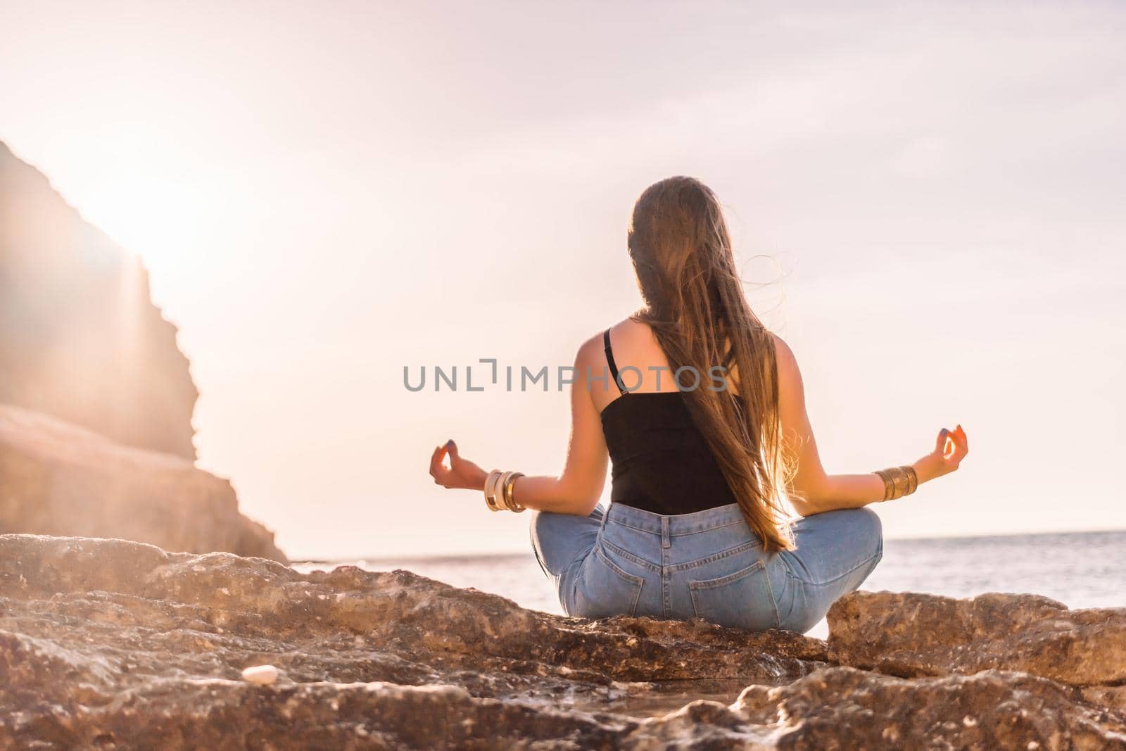 Young woman with long hair in black swimsuit and boho style braclets practicing outdoors on yoga mat by the sea on a sunset. Women's yoga fitness routine. Healthy lifestyle, harmony and meditation by panophotograph