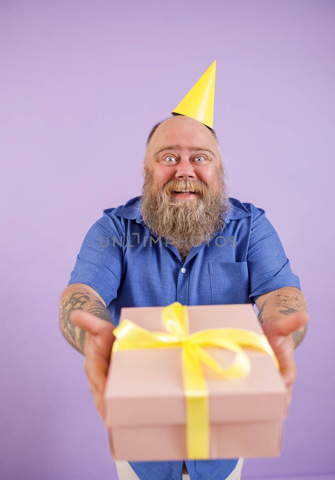 Happy fat man in party hat presents gift box standing on purple background by Yaroslav_astakhov