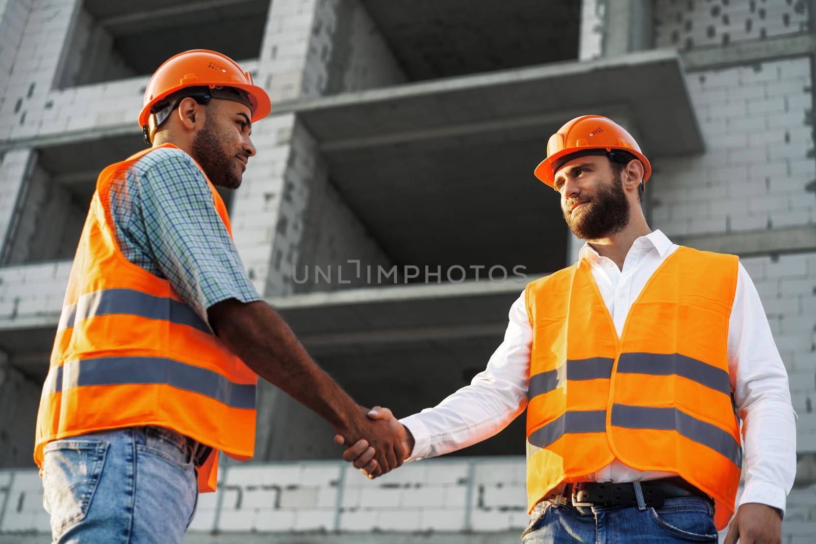 Two men engineers in workwear shaking hands against construction site, close up