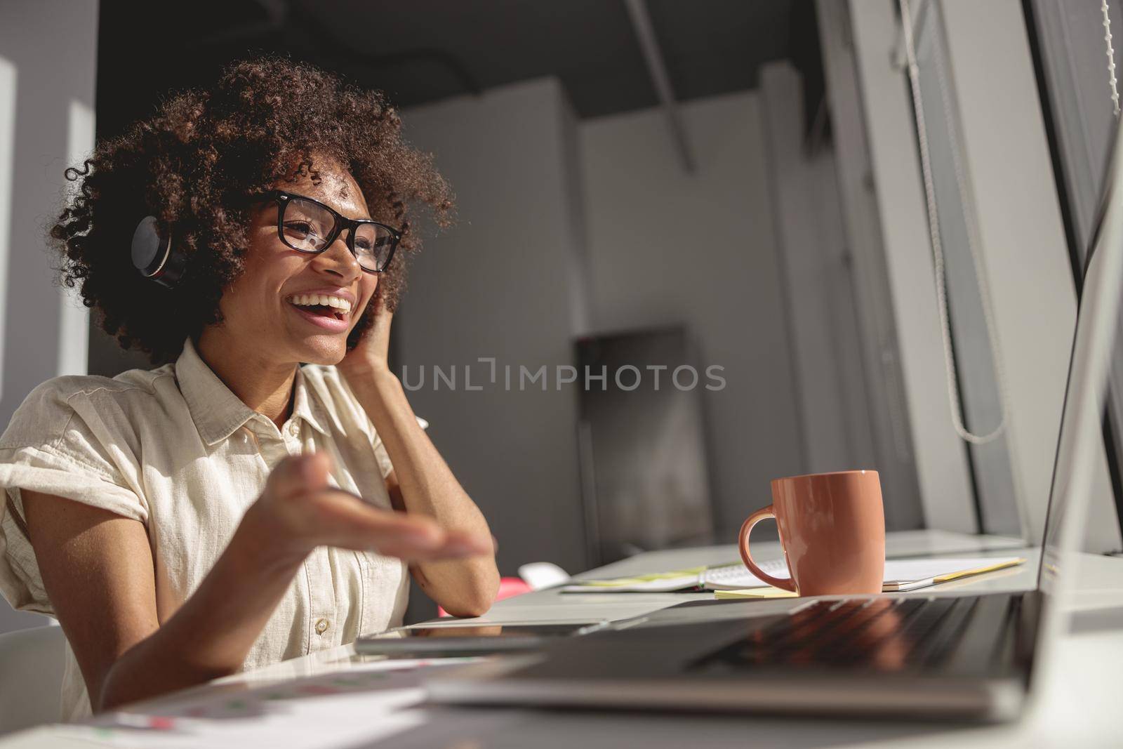 Smiling Afro American lady using headphones and looking at laptop screen while making a video call at workplace