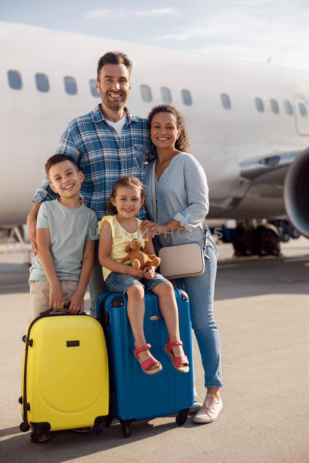 Full length shot of happy family of four standing in front of big airplane on a daytime and smiling at camera. People, traveling, vacation concept