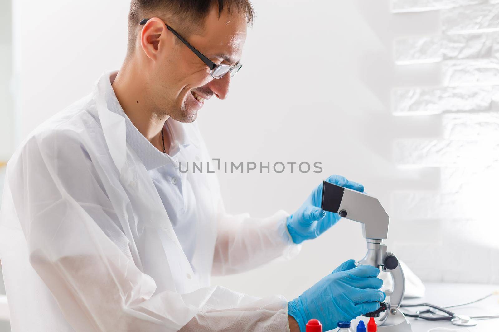 A male doctor or scientist looking through a microscope on a table with laptop computer in background