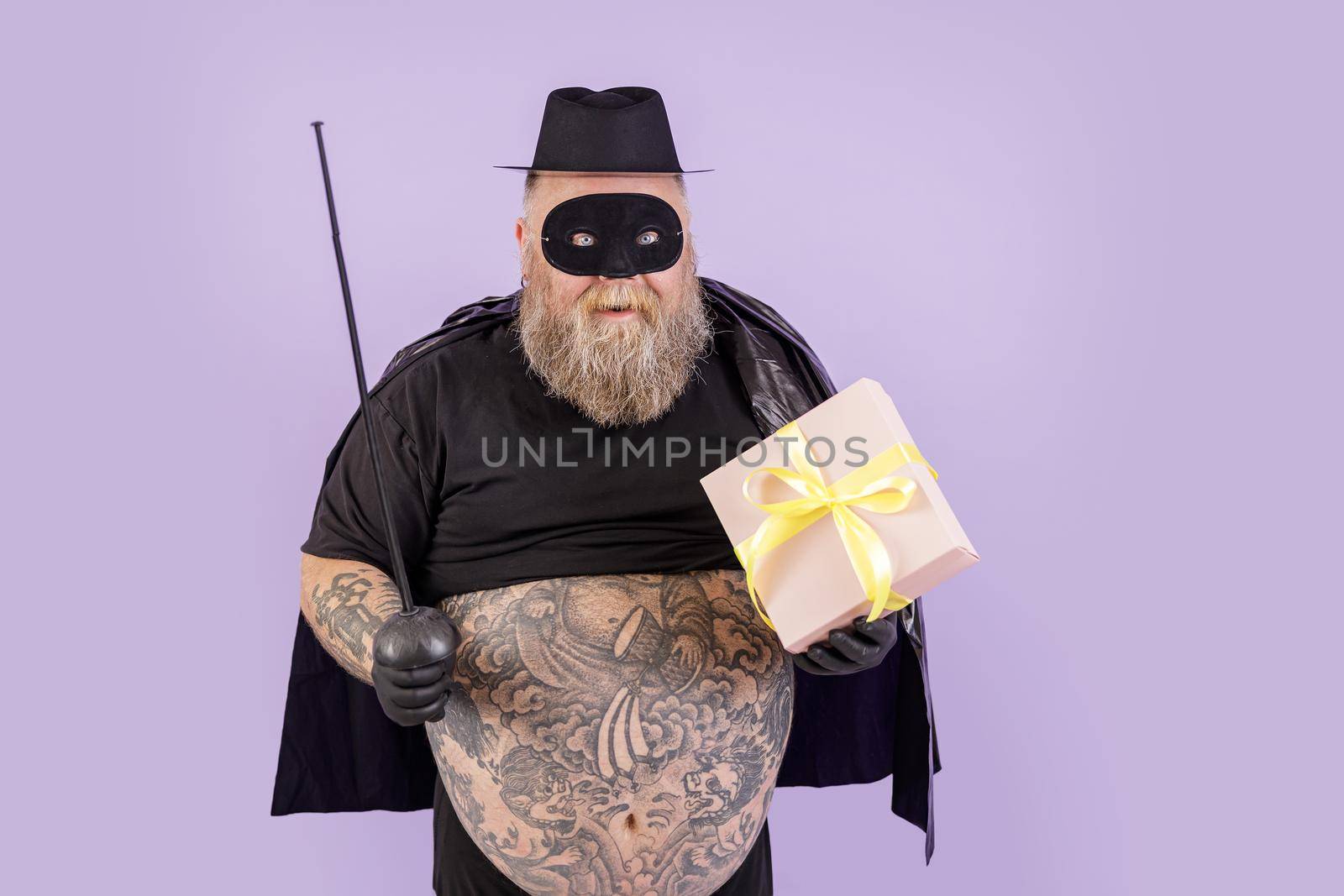 Happy plump gentleman in Zorro suit holds toy epee and gift box on purple background by Yaroslav_astakhov