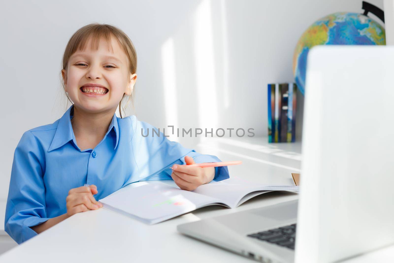 Smart Little Girl Does Homework in Her Living Room. She's Sitting at Her Desk Writes with a Pen in Her Textbooks and Uses Laptop.