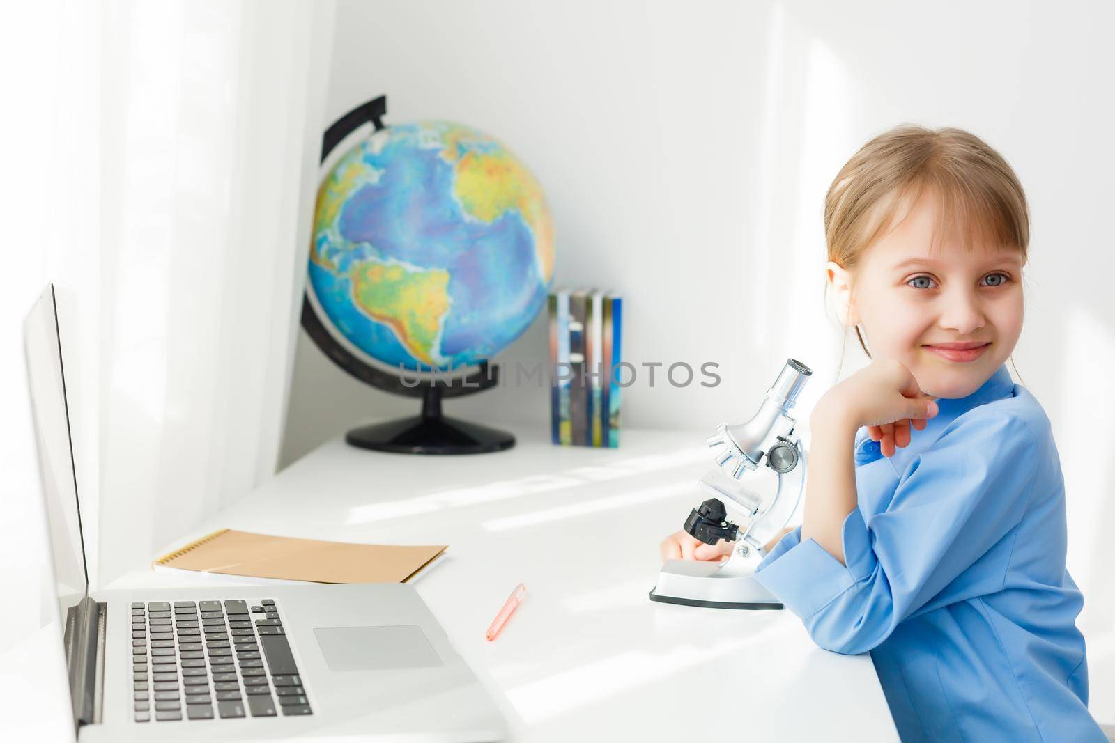 Smart Little Girl Does Homework in Her Living Room. She's Sitting at Her Desk Writes with a Pen in Her Textbooks and Uses Laptop.