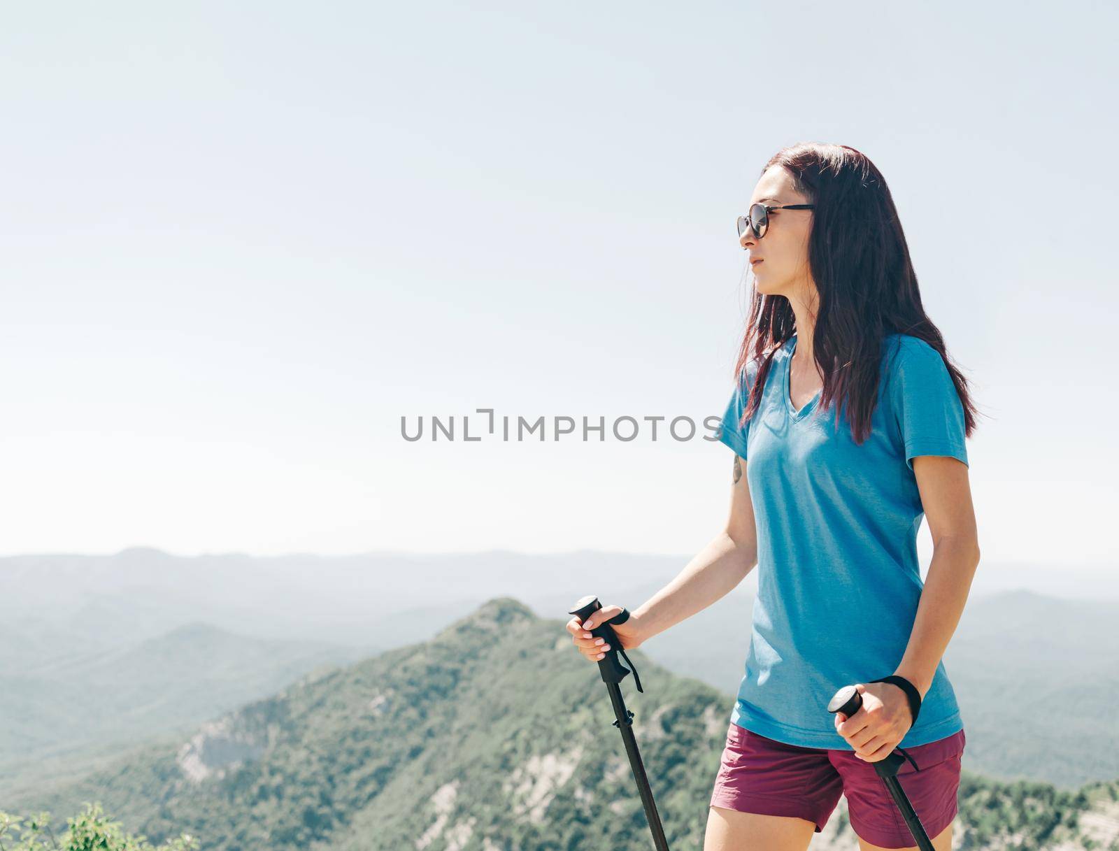 Active sporty young woman in a t-shirt and shorts walking with trekking poles in summer mountains outdoor.