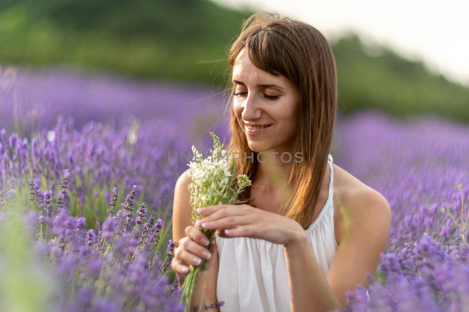 Close up portrait of happy young brunette woman in white dress on blooming fragrant lavender fields with endless rows. Warm sunset light. Bushes of lavender purple aromatic flowers on lavender fields. by panophotograph