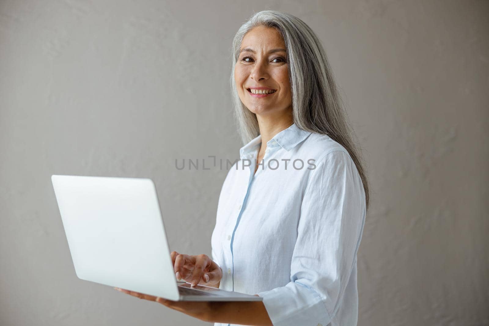 Cheerful silver haired mature Asian businesswoman in white blouse works on modern laptop posing near grey stone wall in studio