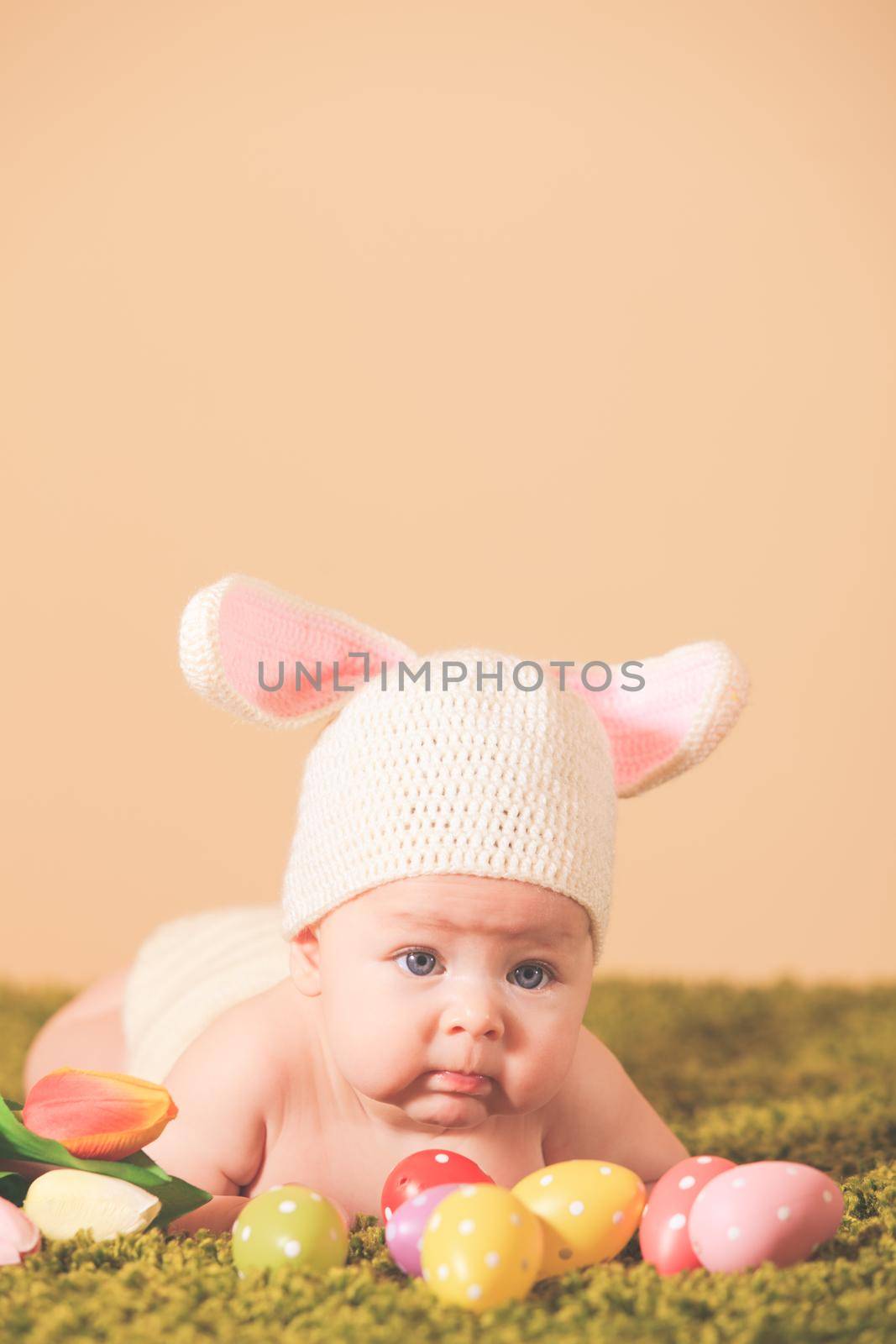 Three months baby lying on his stomach as a Easter bunny on the grass with eggs